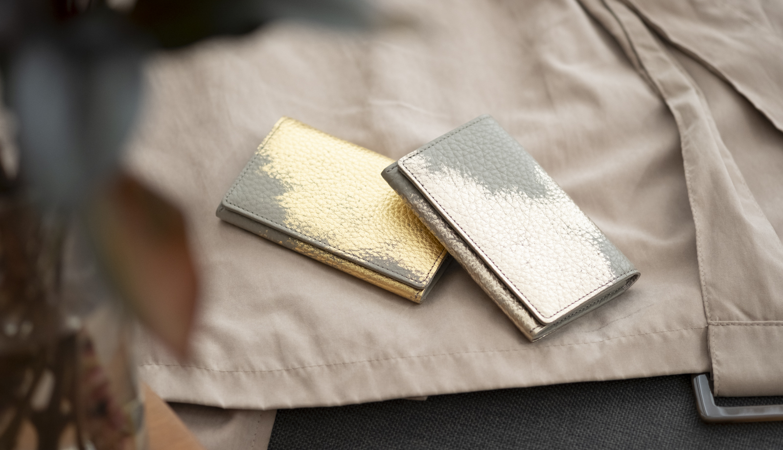 春のトレンチコートの上に静かに置かれた、箔座のレザー名刺入れ。新しい出会いと出発の情景。